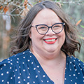 Woman with brown hair wearing black glasses, red lipstick, and a blue polka dot blouse
