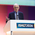 Man with white hair and dark blue round glasses speaks at a podium at ISHLT2026
