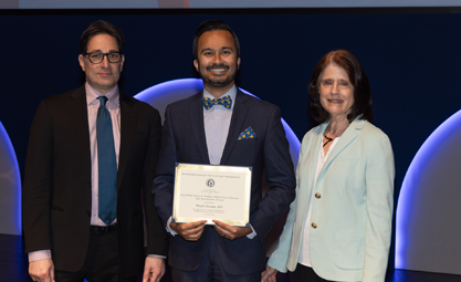 Three people stand next to each other posing for a photo. A white man with black hair wearing a suit stands with a brown man in a suit with a bow tie holding a certificate who stands next to a white woman with brown hair in a light blue suit jacket