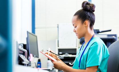 Photo of woman in scrubs sitting at a desk looking at a clipboard