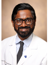 Brown man with black hair and a black beard smiles wearing black frame square glasses and a white doctors coat