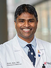 Brown man smiling in a light blue button down shirt, navy blue tie, and white doctor's coat